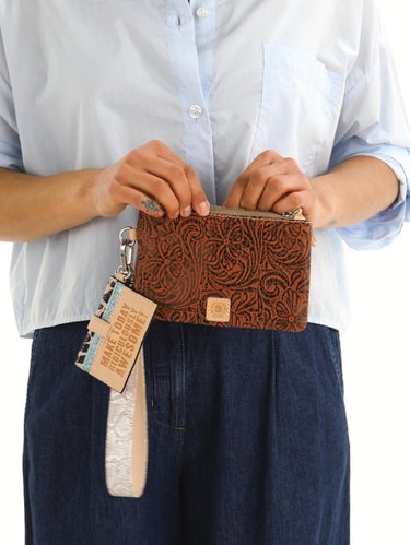 A person wearing a light blue button-up shirt and dark blue jeans holds a brown embossed leather zip pouch with a white wrist strap and a hanging tag that reads 'WELCOME TO MY WORLD'.