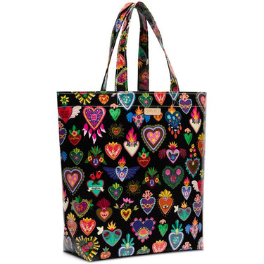 A black tote bag featuring a vibrant, colorful pattern of traditional Mexican Sacred Heart of Jesus motifs, including hearts with wings, flowers, and religious symbols. The bag has two shoulder straps and a small gold-tone brand plaque on the front.