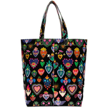 A black tote bag featuring a vibrant, colorful pattern of traditional Mexican Sacred Heart of Jesus motifs, including hearts with flames, roses, and floral details, set against a plain white background.