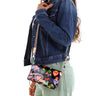 A woman wearing a Consuela black crossbody bag with colorful heart and floral print, tan wristlet strap, and striped woven shoulder strap.