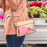 A person wearing a textured peach jacket and blue jeans carries a clear plastic crossbody bag with a beige frame and a pink glittery wallet inside. The bag has a rainbow-striped shoulder strap and a small gold brand plaque that reads 'CONSUELA.
