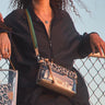 A person wearing a black pinstripe zip-up jacket carries a small crossbody bag with a leopard-print pattern, clear transparent side panels, and a colorful striped strap, photographed outdoors against a fence and sky.