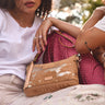 A woman in a white t-shirt holds a tan Consuela crossbody bag with a distressed world map pattern. A bright pink studded purse is also visible in the background.