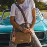 A person wearing a white graphic t-shirt and gray studded jeans sits in front of a teal vintage convertible, carrying a brown embossed crossbody bag with a striped strap.