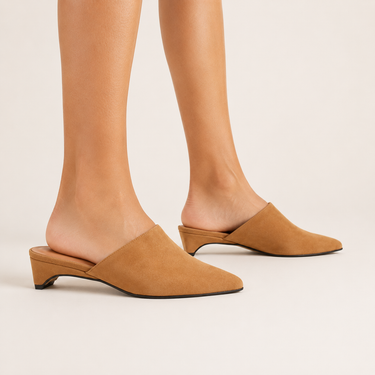 A pair of tan suede mule heels with pointed toes and low block heels, shown on a model's feet against a light beige studio background.