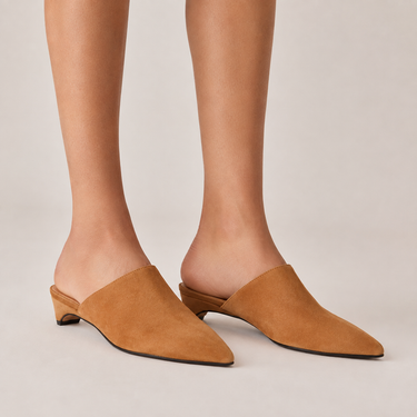 A pair of tan suede mule heels with pointed toes and low block heels, shown on a model's feet against a light beige studio background.