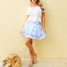 Woman in a white top and blue floral skirt standing next to a dog against a beige wall.