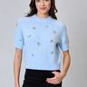 a woman wearing a blue knit short sleeve sweater with sparkling floral embellishments and black pants on a neutral background