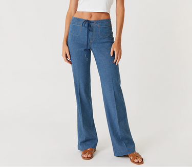 A woman modeling a pair of medium-wash blue wide-leg jeans with a drawstring waist, paired with a white sleeveless crop top and brown sandals, against a plain light gray studio background.