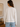 Person wearing a beige sweater and blue jeans in a room with a vase and framed picture on a shelf.