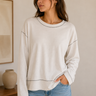Woman wearing a beige sweatshirt and blue jeans in a room with a vase and painting on a shelf.
