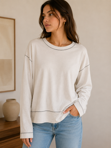 Woman wearing a beige sweatshirt and blue jeans in a room with a vase and painting on a shelf.