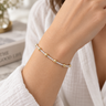 A delicate gold and silver beaded bracelet worn on a woman's wrist, styled with a white textured robe in a bright, softly blurred indoor setting.