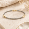 A beaded stretch bracelet featuring gray stone beads and polished gold spacer beads, displayed on a textured beige stone surface with neutral fabric and dried flowers in the background.