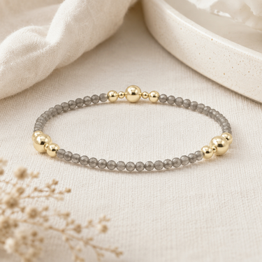 A delicate beaded bracelet with gray translucent round beads and polished gold spacer beads, displayed on a textured beige fabric surface.