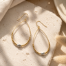 A pair of gold teardrop hoop earrings with a beaded lower section and a small silver oval bead, displayed on a textured beige stone with soft natural shadows.