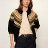 A woman modeling a black knit zip-up jacket with a tan and beige geometric pattern across the shoulders and upper chest, paired with a cream t-shirt and dark blue jeans against a plain light background.