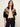 A woman modeling a black knit zip-up jacket with a tan and beige geometric pattern across the shoulders and upper chest, paired with a cream t-shirt and dark blue jeans against a plain light background.