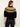 A woman shown from the back wearing a black knit sweater with a brown and beige geometric Fair Isle yoke pattern, paired with dark blue jeans, against a plain off-white background.