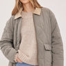 A woman wearing a quilted taupe-gray jacket with a beige collar, layered over a beige knit sweater and blue jeans, against a light gray studio background.