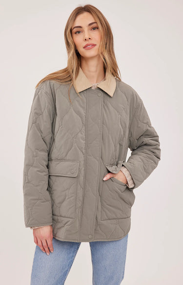 A woman modeling a light gray quilted jacket with a beige collar, worn over a beige sweater and blue jeans against a plain light gray studio background.