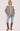 A woman modeling a light gray quilted jacket with a beige collar, worn over a beige sweater, blue jeans, and brown knee-high boots, against a plain light gray studio background.
