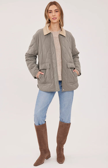 A woman modeling a light gray quilted jacket with a beige collar, worn over a beige sweater, blue jeans, and brown knee-high boots, against a plain light gray studio background.