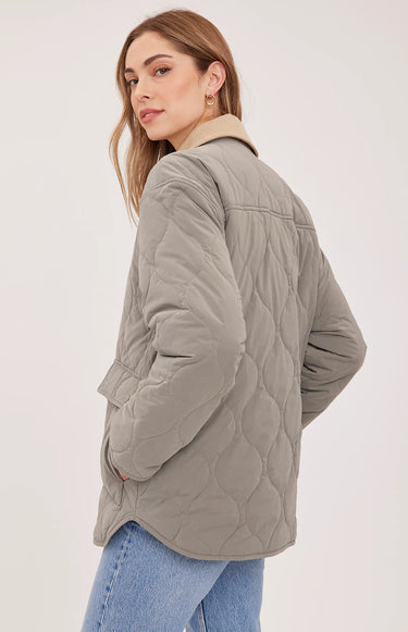 A woman modeling a light gray quilted jacket with a beige collar, worn over a beige sweater and blue jeans against a plain light gray studio background.