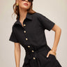 Woman wearing a black romper with gold buttons against a beige background
