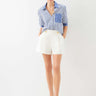 A woman modeling a blue and white vertically striped button-down shirt with rolled sleeves and white shorts, standing against a plain white studio background.