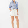 A woman modeling a blue and white vertically striped button-down shirt with rolled sleeves, paired with white shorts and beige heeled sandals, against a plain white studio background.