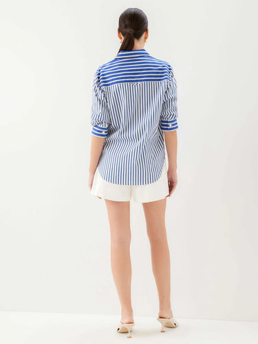 A woman viewed from the back wearing a blue and white vertically striped button-up shirt with white shorts and beige heels, set against a plain white studio background.