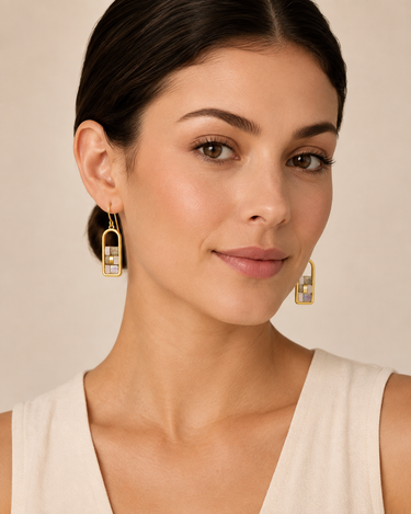 A close-up of a woman wearing gold geometric drop earrings with clear rectangular stones, set against a soft beige background.