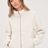 A woman wearing a cream sherpa fleece zip-up jacket over dark blue jeans, photographed against a neutral light gray studio background.