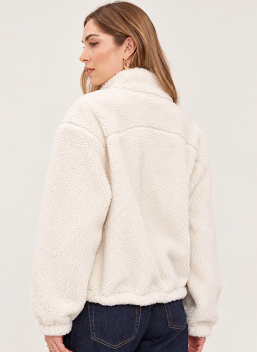 A woman shown from the back wearing a cream sherpa fleece jacket and dark blue jeans against a plain light gray studio background.