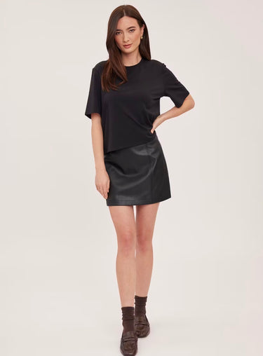 A woman wearing a black tee, faux leather skirt in black with brown loafers on a white background.