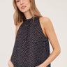 A woman wearing a sleeveless black and white polka dot flowy blouse with a mock neck and black jeans on a white background.