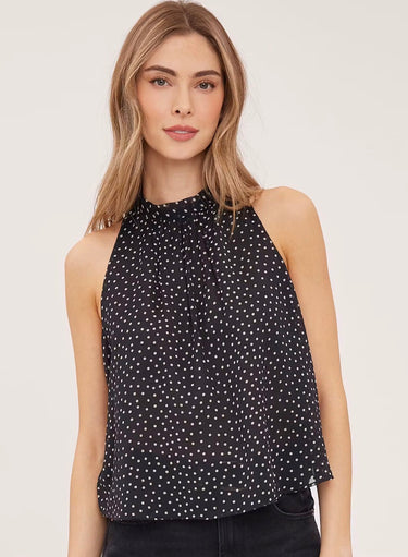  A woman wearing a sleeveless black and white polka dot flowy blouse with a mock neck and black jeans on a white background.