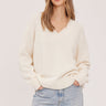 A woman wearing a cream v-neck pullover and blue jeans on a white background