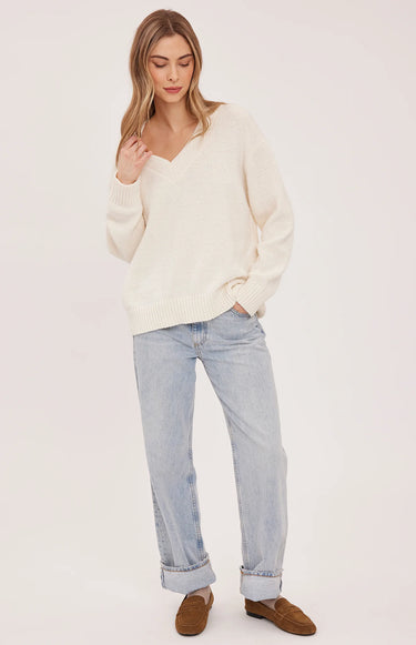 A woman wearing a cream v-neck pullover and blue jeans on a white background
