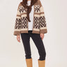 A woman modeling a brown and beige patterned knit cardigan with a matching brown scarf, paired with black pants and tan boots, against a light beige studio background.