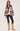A woman modeling a brown and beige patterned knit cardigan with a matching brown scarf, paired with black pants and tan boots, against a light beige studio background.