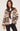 A woman wearing a brown, cream, and dark brown knit sweater with geometric patterns, shown from a three-quarters back view against a light beige studio background.