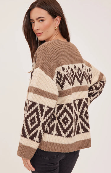 A woman wearing a brown, cream, and dark brown knit sweater with geometric patterns, shown from a three-quarters back view against a light beige studio background.