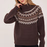 A woman wearing  a brown turtle neck with a fair isle print and black  jeans on a white background.