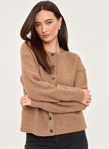 A woman wearing a textured knit button-front cardigan in a warm camel brown color, paired with black pants, against a plain light gray studio background.