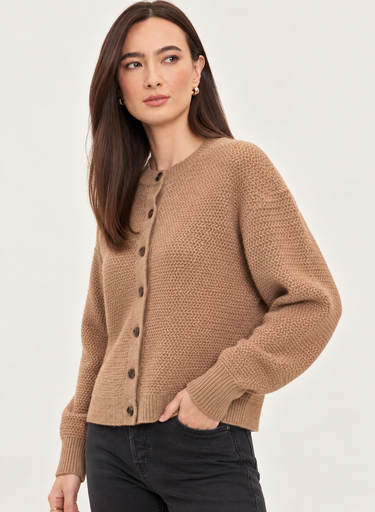 A woman wearing a textured knit cardigan sweater in a warm camel brown color, paired with black jeans, against a plain light gray studio background.
