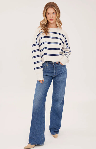 A woman wearing a cream and navy stripe oversized sweater and blue jeans on a white background