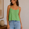 Woman wearing a green lace top and blue jeans in a room with a wooden shelf and framed picture.
