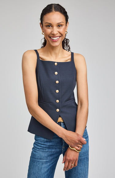 Woman wearing a navy blue sleeveless top with buttons and jeans on a gray background